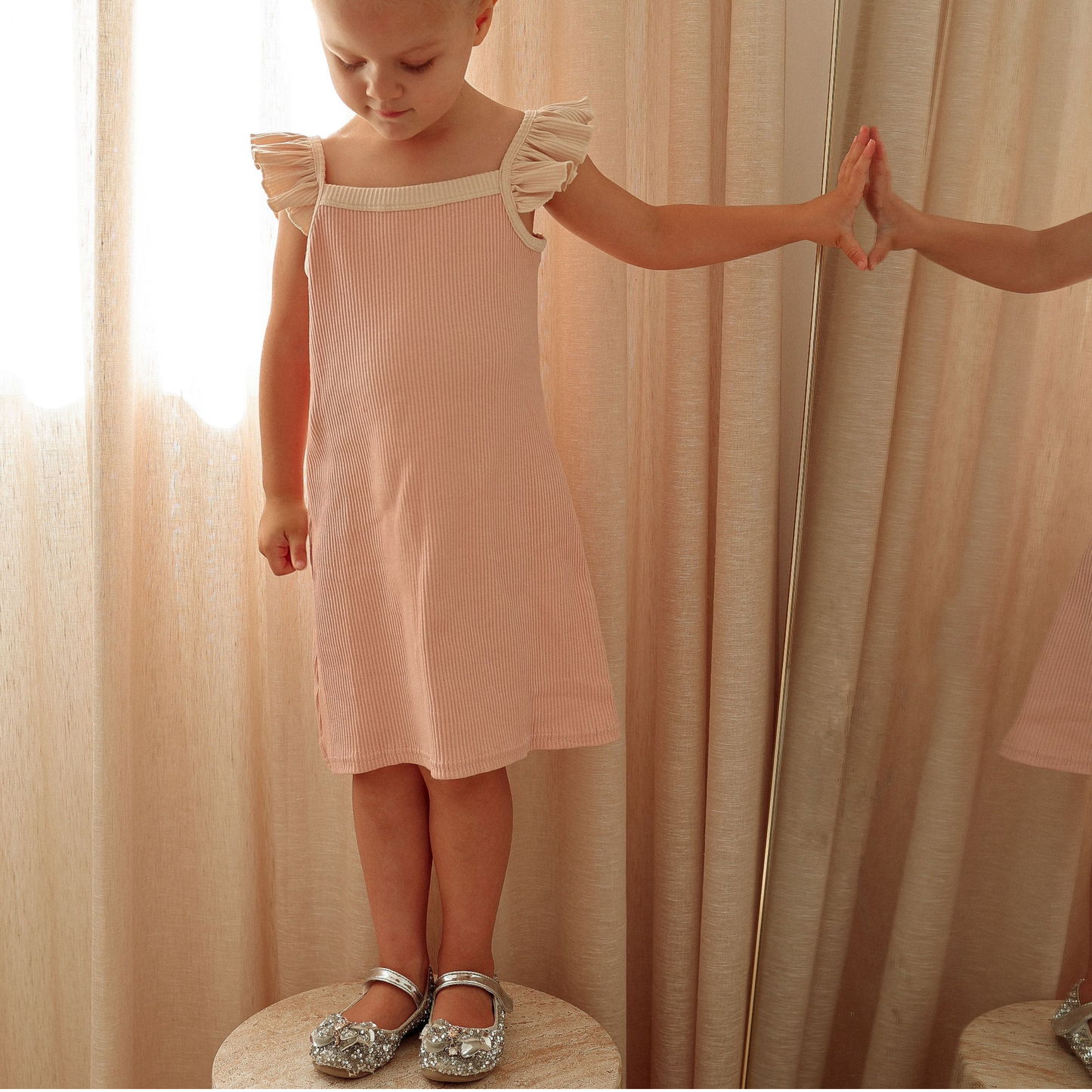 Young girl in a pink dress standing next to a mirror with a blurred reflection.