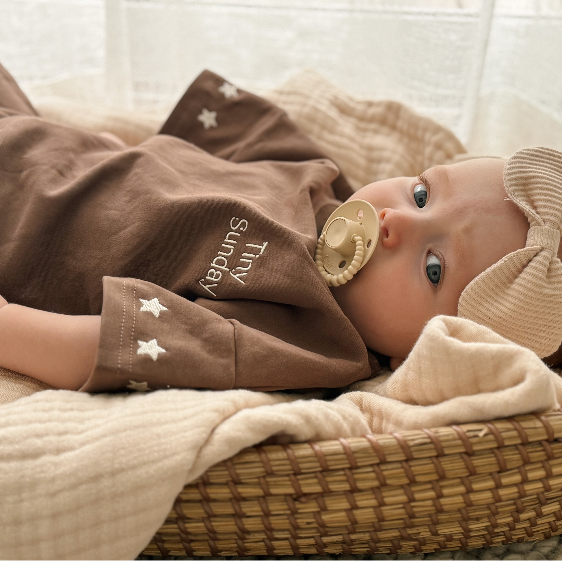 Baby lying in a basket with brown outfit and dummy, surrounded by soft textures.