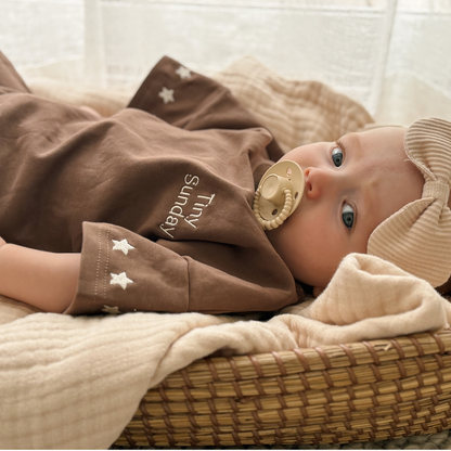Baby lying in a basket with brown outfit and dummy, surrounded by soft textures.