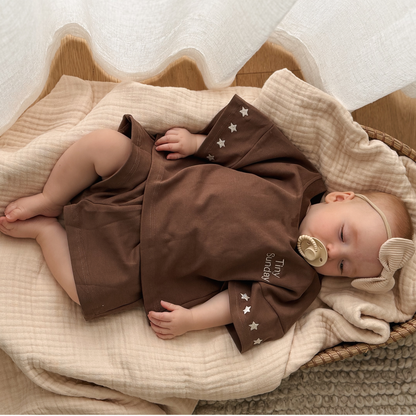 Baby in a brown outfit with star patterns lying on a beige blanket.